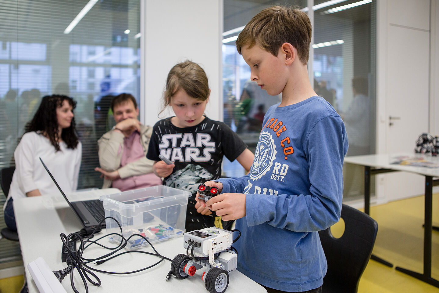 Julius bastelt an einem Roboter herum, während seine Tischnachberin am Laptop programmiert. Seine Eltern sitzen im Hintergrund und begutachten die Arbeit.