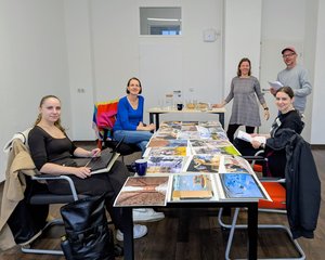 vier Frauen und ein Mann schauen zahlreiche Bilder an, die auf einem Tisch liegen