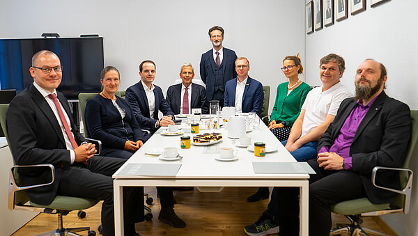 mehrere Personen sitzen als Gruppe an einem Tisch und lächeln in die Kamera
