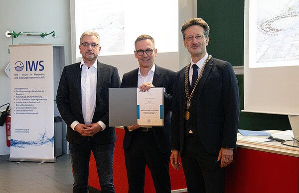 drei Männer in Anzügen stehen zusammen, der Mittlere hät eine Urkunde hoch