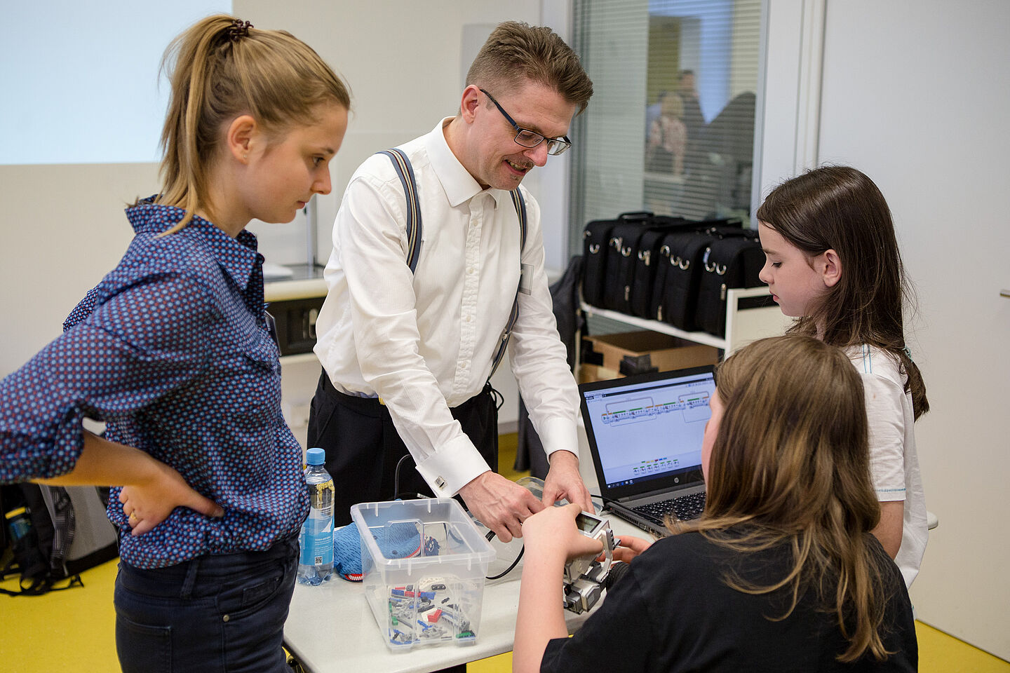 Zwei Kinder schauen etwas ratlos auf den Roboter, den eins von ihnen in der Hand hält. Herr Prof. Wagner steht an der anderen Seite des Tisches und hilft mit gezielten Handgriffen.