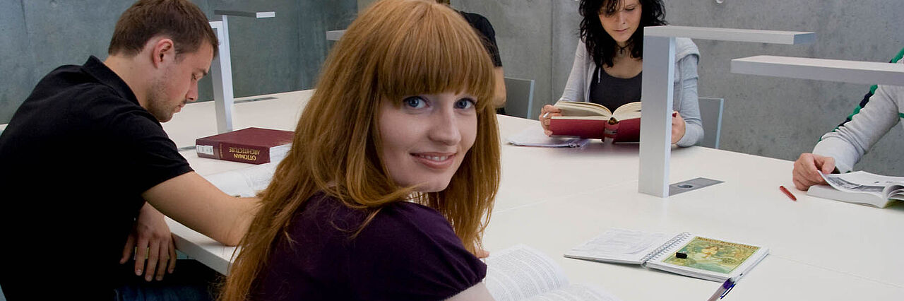 Studierende sitzen an einem Tisch in der Bibliothek in der HTWK. Die Studentin im Vordergrund ist zu Kamera hingewandt und lächelt.