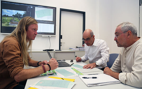 HTWK-Mitarbeiter Felix Oertel, Prof. Ralf Thiele und Prof. Ronald Scherzer-Heidenberger besprechen die Messergebnisse der geotechnischen Untersuchungen am Tisch sitzend. Im Hintergrund sind auf einem großen Bildschirm Pläne und Fotos zu sehen.