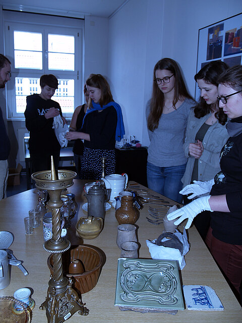 Übungen an Objekten Auf einem Tisch stehen Objekte, dahinter Menschen