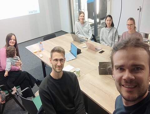 Es sitzen mehrere Studierenden an einem Tisch. Eine Person macht ein Selfie-Bild von der Gruppe, während alle in die Kamera lächeln.