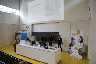 AM³-Symposium Vier Personen stehen am Rednerpult im Hörsaal. An die Wand wird eine Folie projiziert, und eine Frau spricht in ein Mikrofon