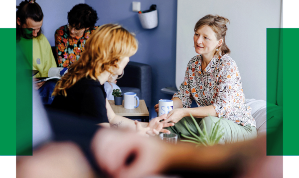 Gespräch in gemütlicher Runde Fünf Personen sitzen in einem hellen Raum, unterhalten sich und trinken Kaffee. Zwei Frauen sprechen sich direkt gegenüber.