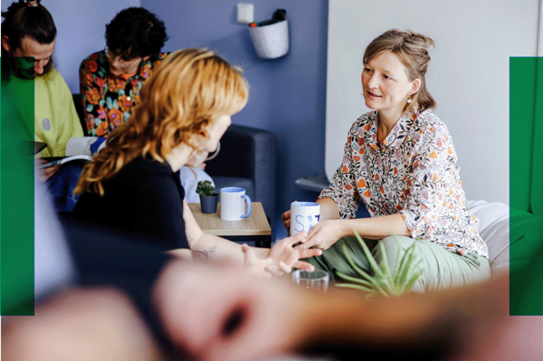 Fünf Personen sitzen in einem hellen Raum, unterhalten sich und trinken Kaffee. Zwei Frauen sprechen sich direkt gegenüber.