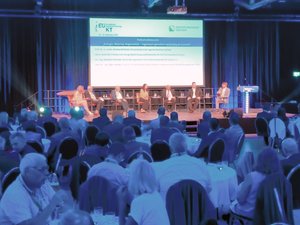 mehrere Personen sitzen auf einer Bühne und diskutieren, im Saal sind viele Besucher an Tischen