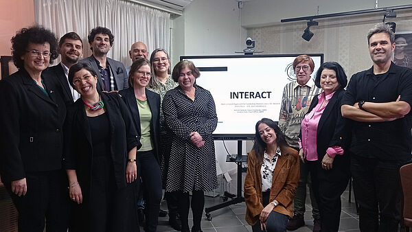 Das Foto zeigt 12 Frauen und Männer stehend vor einem Bildschirm mit der Aufschrift INTERACT