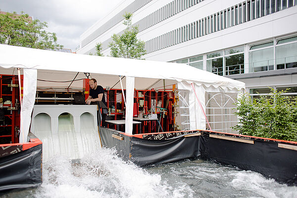 Unter einem langen Pavillon sind im Freien viele Geräte des Wasserbaulabors aufgebaut. In der Mitte ist eine Talsperre errichtet, durch die Wasser fließt