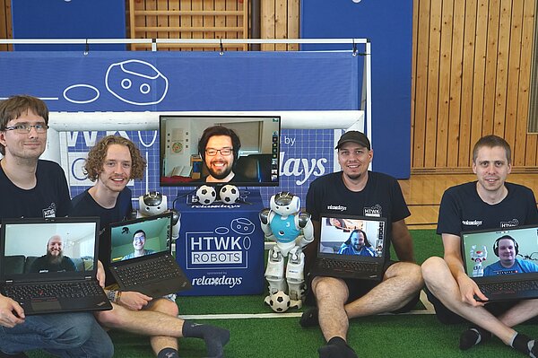 Teammitglieder (in Präsenz, v.l.n.r.): Tobias Jagla, Felix Loos, Stephan Bischoff und Jörg Schließer. Auf den Laptops zu sehen sind Teammitglieder, die von Ferne unterstützt haben (v.l.n.r.): Teamchef Rico Tilgner, Erik Behrendt, Tobias Kalbitz (groß Vier Männer mit Laptops auf dem Schoß vor kleinem Tor und mit humanoidem Roboter