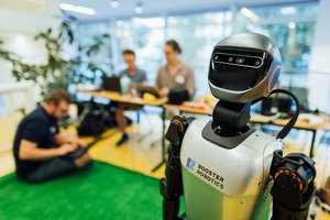 "Booster". (Foto: Christian Hüller/HTWK Leipzig) Roboter im Vordergrund, dahinter sitzen Menschen auf Kunstrasen mit Laptops