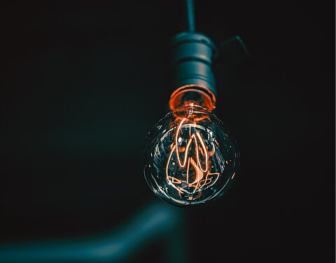 Eine schwarze Glühbirne mit einem sehr langen Leuchtdraht hängt vor einem schwarzen Hintergrund.