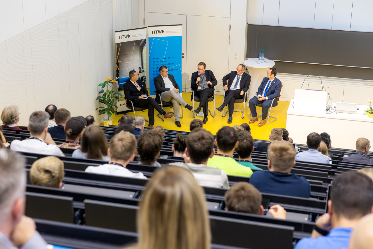 Blick von oben nach unten im Hörsaal, ganz hinten 5 Personen der Podiumsdiskussionen zu sehen, Hörsaal Sitzreichen von hinten im Vordergrund zu sehen 