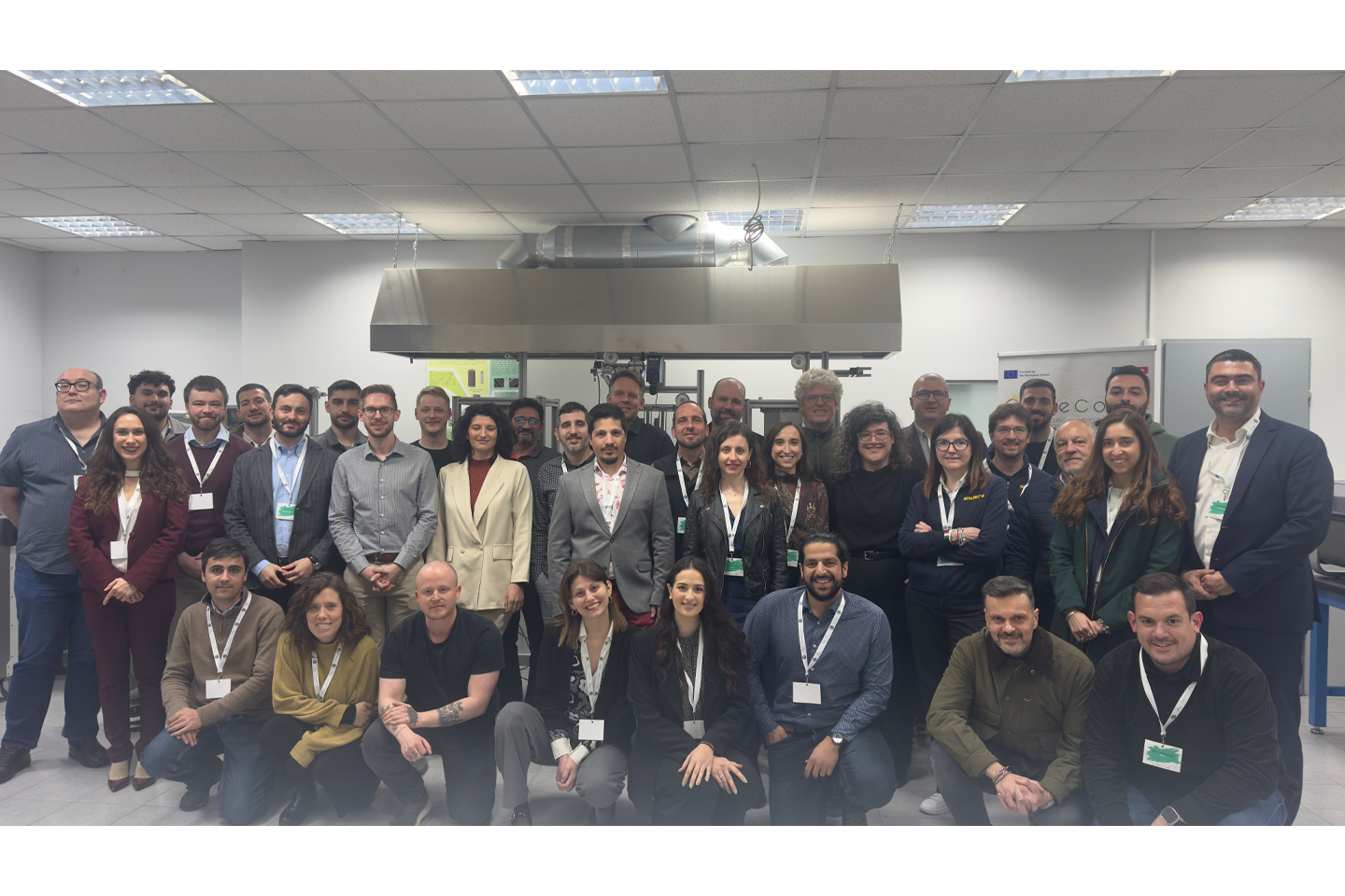 Sehr große Gruppe an Menschen posieren für ein Gruppenfoto und schauen in die Kamera, im Hintergrund Neonröhren und großer Lüfter an der Decke