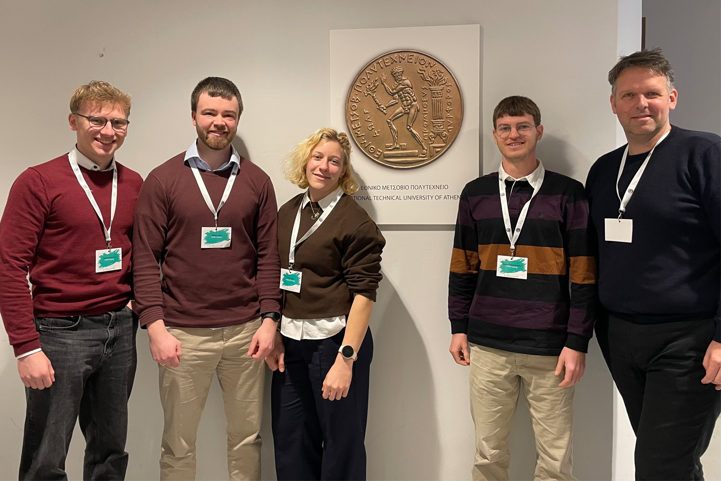 Fünf Personen schauen in die Kamera, im Hintergrund goldenes Emblem an der Wand